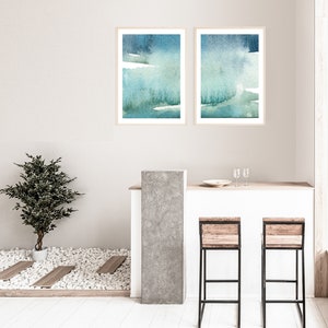 May include: Two framed abstract watercolour paintings in shades of blue and green, hanging on a white wall above a bar counter with two wooden bar stools.