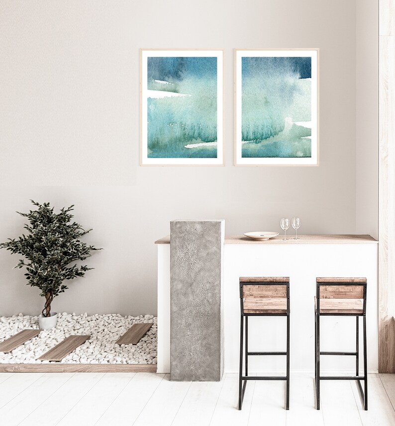 May include: Two framed abstract watercolour paintings in shades of blue and green, hanging on a white wall above a bar counter with two wooden bar stools.