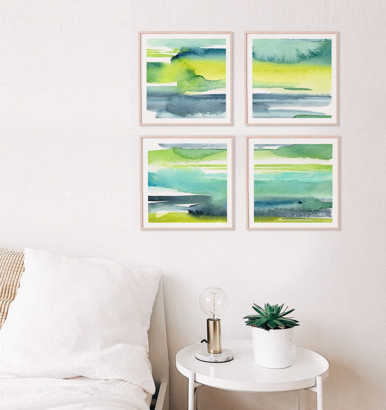 May include: Set of four abstract watercolor paintings in shades of green, blue, and yellow. The paintings are framed in a light wood frame and are hung on a white wall.