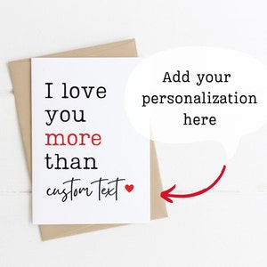 May include: A blank card with the text "I love you more than" followed by a space for personalization. The text "custom text" is written in cursive with a red heart next to it. The card is on a white wooden background.
