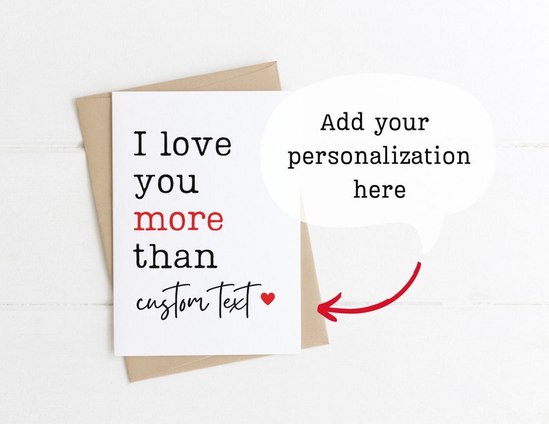 May include: A blank card with the text "I love you more than" followed by a space for personalization. The text "custom text" is written in cursive with a red heart next to it. The card is on a white wooden background.