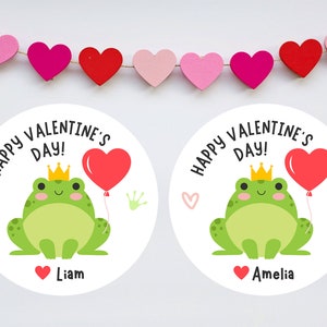 May include: Two round white stickers with a green frog holding a red heart balloon. The stickers say "Happy Valentine's Day!" and have a small heart with the name "Liam" or "Amelia" on the bottom.