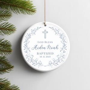 Może przedstawiać: Okrągła, biała ceramiczna ozdoba z niebieskim krzyżem i wieńcem kwiatowym. Tekst brzmi "GOD BLESS Aiden Noah BAPTIZED 10. 11. 2025". Do zawieszenia przymocowany jest brązowy sznurek. Przedmiot dekoracyjny.