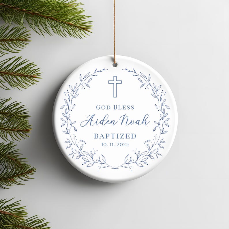 Może przedstawiać: Okrągła, biała ceramiczna ozdoba z niebieskim krzyżem i wieńcem kwiatowym. Tekst brzmi "GOD BLESS Aiden Noah BAPTIZED 10. 11. 2025". Do zawieszenia przymocowany jest brązowy sznurek. Przedmiot dekoracyjny.