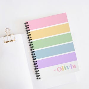 Op de afbeelding: Een spiraalgebonden notitieboek met een pastelkleurige regenboog gestreepte omslag. De omslag heeft roze, gele, groene, blauwe en paarse strepen. De naam "Olivia" is in pastelkleuren gedrukt met een klein hartje. Een gouden paperclip is zichtbaar.