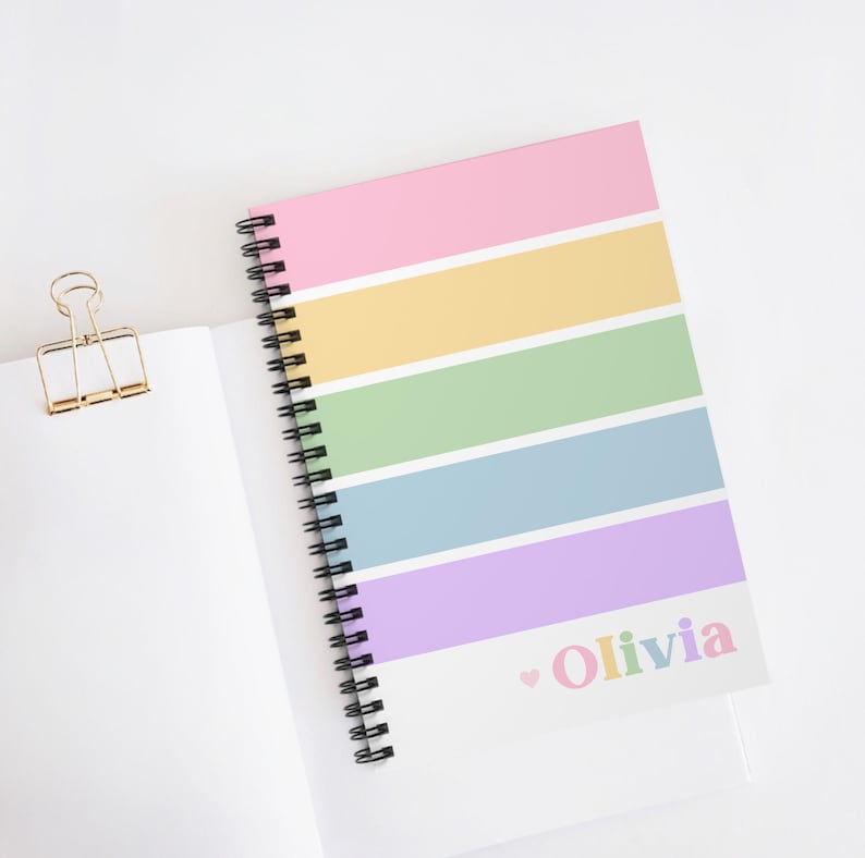 Op de afbeelding: Een spiraalgebonden notitieboek met een pastelkleurige regenboog gestreepte omslag. De omslag heeft roze, gele, groene, blauwe en paarse strepen. De naam "Olivia" is in pastelkleuren gedrukt met een klein hartje. Een gouden paperclip is zichtbaar.