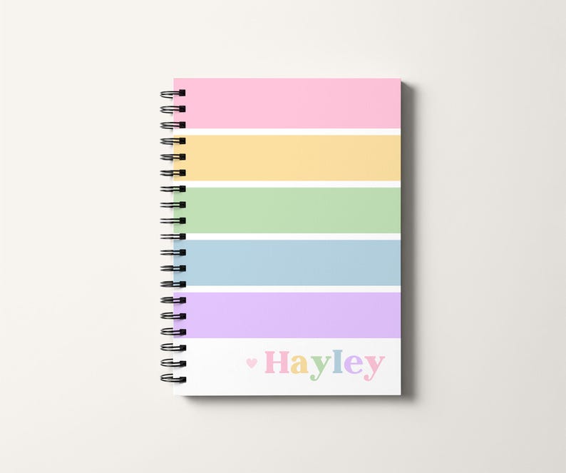 Op de afbeelding: Een spiraalgebonden notitieboek met een pastelkleurige regenboog gestreepte omslag. De omslag heeft horizontale strepen in roze, geel, groen, blauw en paars. De naam "Hayley" staat in regenboogkleuren onderaan.