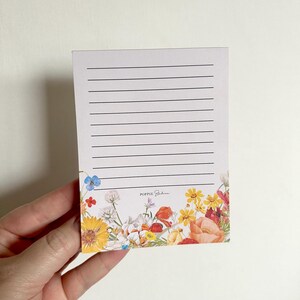 May include: A white notepad with lined paper and a floral border. The border features a variety of flowers in shades of red, orange, yellow, blue, and green. The text "POPPIE Studio" is printed in black at the bottom of the notepad.