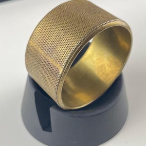 May include: A gold-colored, wide bangle bracelet with a perforated design. The bracelet is resting on a black stand, showcasing its texture and shape. The bangle's surface has a pattern of small holes.