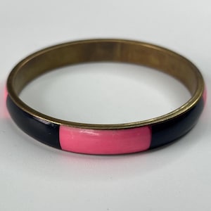 May include: A brass bangle bracelet with a black and pink enamel design. The bracelet has a circular shape and a smooth, polished finish. The black and pink sections alternate around the circumference of the bracelet.