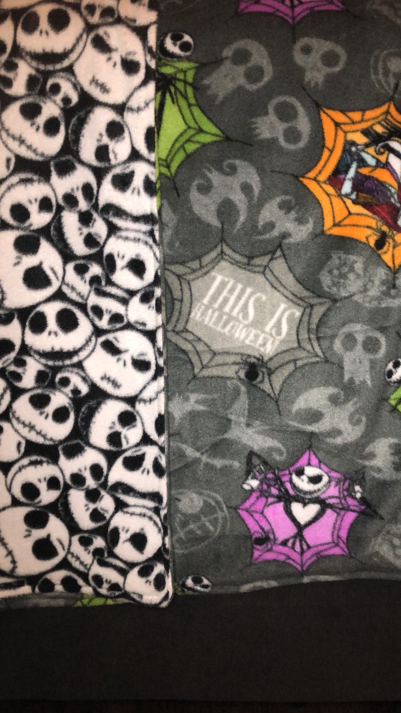 May include: A fleece blanket featuring a patchwork design. One side displays a pattern of Jack Skellington faces in black and white. The other side has a gray background with spiderwebs, skulls, and the text "THIS IS HALLOWEEN".