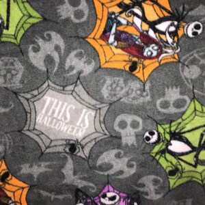 May include: A gray fabric with a repeating pattern of skulls, spiderwebs, and characters from The Nightmare Before Christmas. The text "THIS IS HALLOWEEN" is visible. The spiderwebs are in orange, green, and purple.