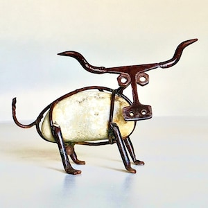 May include: A small, whimsical sculpture of a bull made from metal and a white rock. The bull has long horns, a simple face, and a rounded body.