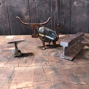 May include: A metal sculpture of a bull made from wire and a stone. The sculpture is sitting on a wooden surface with a small metal anvil in the foreground.