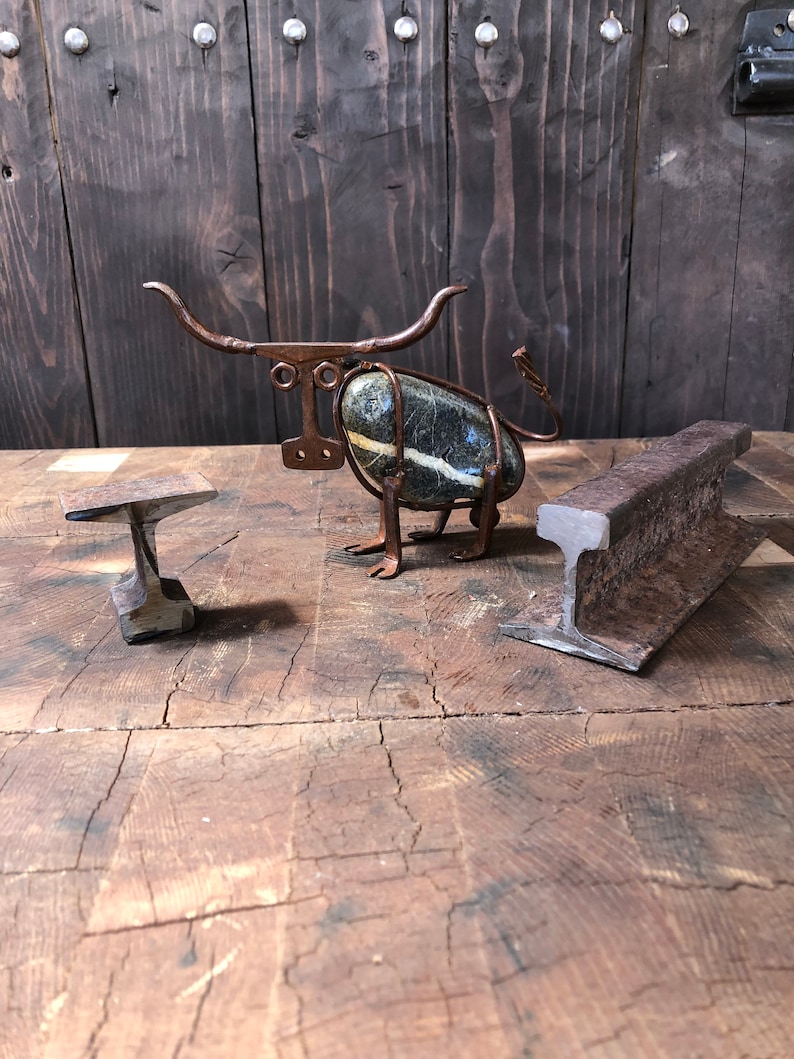 May include: A metal sculpture of a bull made from wire and a stone. The sculpture is sitting on a wooden surface with a small metal anvil in the foreground.