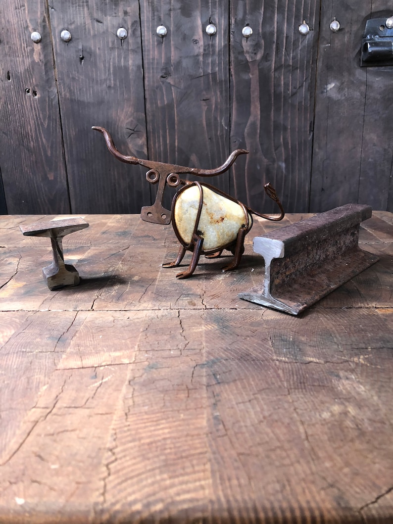 May include: A metal sculpture of a bull made from scrap metal and a white rock. The sculpture is sitting on a wooden table with a metal anvil and a railway track.
