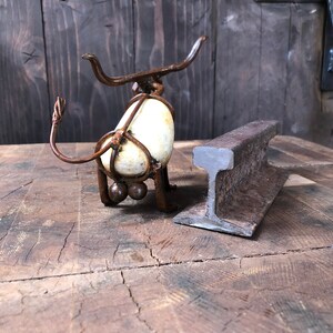 May include: A rusty metal sculpture of a bull with a white rock for a body. The bull is standing on a wooden surface next to a rusty metal railway track.