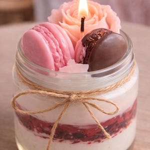Puede incluir: Una vela decorativa en un tarro de cristal transparente, con una mecha encendida, una rosa rosa y dos piezas de cera con forma de macaron. El tarro está atado con hilo y relleno con capas de cera blanca y roja.