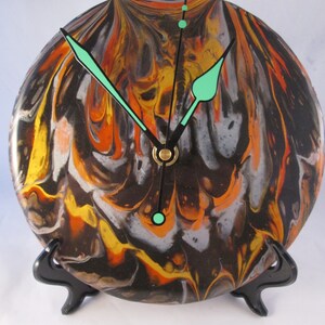 May include: A round clock with a black, orange, and white marbled design. The clock has black hands with green accents and a gold center. The clock is on a black stand.
