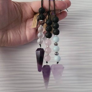May include: Three amethyst crystal pendulums with black lava rock and gemstone beads. The pendulums are attached to black cords. The beads are clear, pale pink, and light blue.