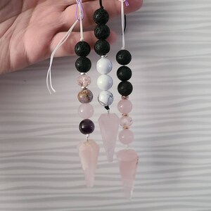 May include: Three pink crystal pendulums with black lava rock beads, white and brown beads, and amethyst beads. The pendulums are hanging from strings.
