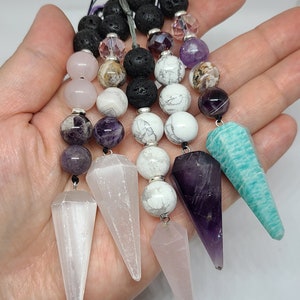 May include: A set of six crystal pendulums with various colors and shapes, including amethyst, selenite, rose quartz, and aventurine. The pendulums are attached to beaded chains with lava rock, white and pink beads.