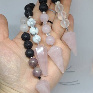 May include: Three pink crystal point pendulums with black lava rock, white howlite, and clear crystal beads on a grey cord.