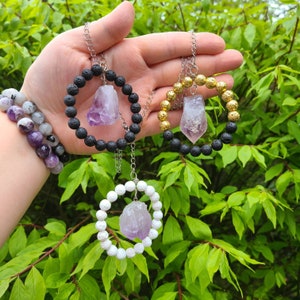 May include: Three necklaces with amethyst crystal pendants. Each necklace has a different style of bead: black lava rock, white beads, and gold beads.