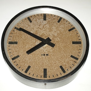 Può includere: Un orologio rotondo bianco e nero con un quadrante marrone e lancette nere. L'orologio ha un bordo argentato e il testo "IBM" è stampato sul quadrante.