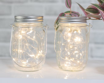 Puede incluir: Dos tarros de vidrio transparente con tapas plateadas y asas de alambre, llenos de luces blancas de hadas. Los tarros están sobre una superficie blanca con una planta verde en el fondo.