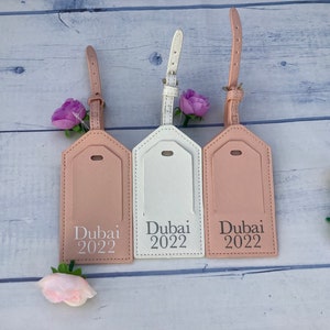 Puede incluir: Tres etiquetas de equipaje con acabado de cuero rosa, blanco y beige. Cada etiqueta tiene una inscripci&oacute;n plateada que dice "Dubai 2022".