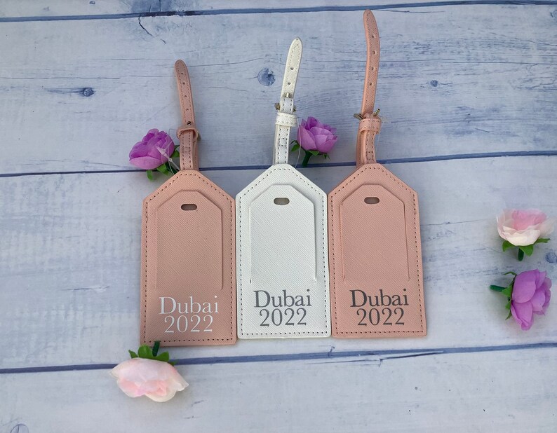 Puede incluir: Tres etiquetas de equipaje con acabado de cuero rosa, blanco y beige. Cada etiqueta tiene una inscripci&oacute;n plateada que dice "Dubai 2022".