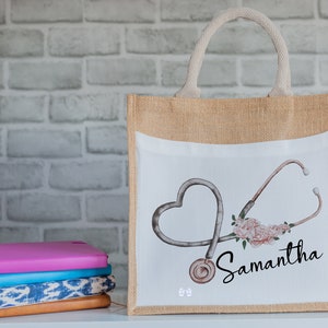 K&ouml;nnte beinhalten: Eine beige Jute-Tragetasche mit einem wei&szlig;en Canvas-Einsatz, der ein Stethoskop in Herzform und rosa Blumen zeigt. Der Name "Samantha" ist in schwarzer Kursivschrift auf der Tasche geschrieben.