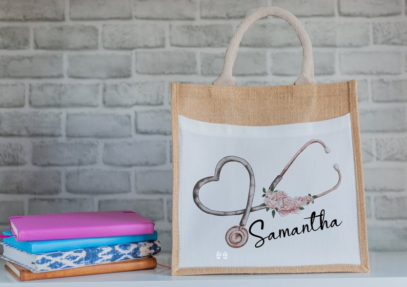 K&ouml;nnte beinhalten: Eine beige Jute-Tragetasche mit einem wei&szlig;en Canvas-Einsatz, der ein Stethoskop in Herzform und rosa Blumen zeigt. Der Name "Samantha" ist in schwarzer Kursivschrift auf der Tasche geschrieben.