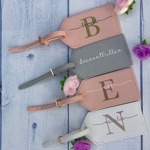 Pu&ograve; includere: Quattro etichette per bagagli personalizzate con cinturini in pelle. Le etichette sono rosa, grigie, bianche e rosa chiaro. Ogni etichetta ha una lettera dorata o argentata e un nome o un'iniziale.