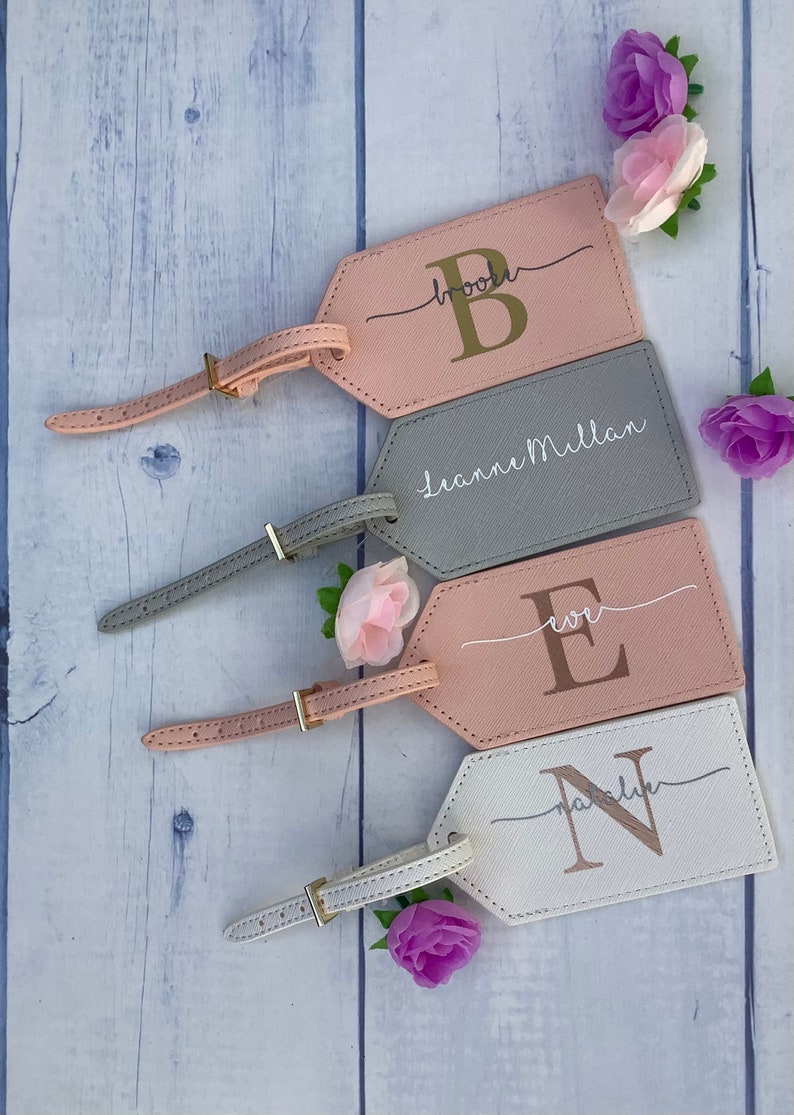 Pu&ograve; includere: Quattro etichette per bagagli personalizzate con cinturini in pelle. Le etichette sono rosa, grigie, bianche e rosa chiaro. Ogni etichetta ha una lettera dorata o argentata e un nome o un'iniziale.