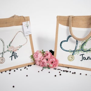 K&ouml;nnte beinhalten: Zwei Jute-Tragetaschen mit wei&szlig;en Stoff-Eins&auml;tzen. Die Taschen zeigen ein Stethoskop mit einem Herz-Design und floralen Akzenten. Die Taschen sind mit den Namen "Ali" und "Jane" personalisiert.