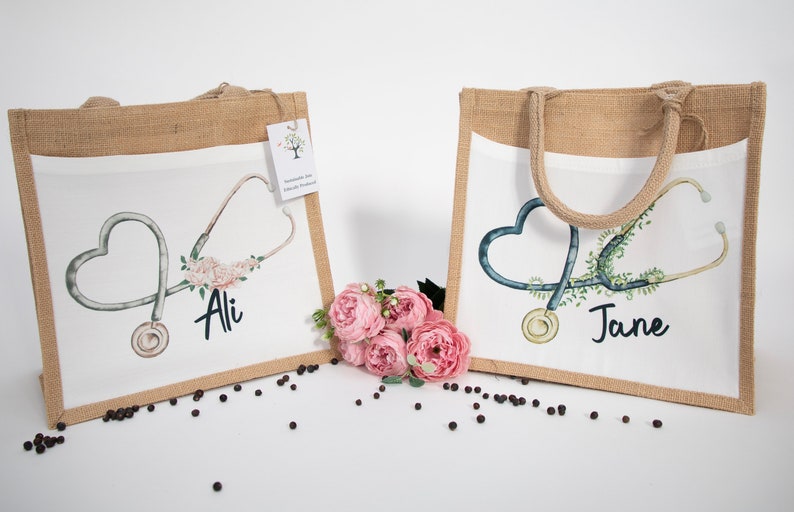 K&ouml;nnte beinhalten: Zwei Jute-Tragetaschen mit wei&szlig;en Stoff-Eins&auml;tzen. Die Taschen zeigen ein Stethoskop mit einem Herz-Design und floralen Akzenten. Die Taschen sind mit den Namen "Ali" und "Jane" personalisiert.