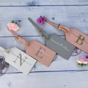 Pu&ograve; includere: Quattro etichette per bagagli personalizzate con nomi e iniziali diversi. Le etichette sono realizzate in un materiale morbido, rosa chiaro e grigio. Le iniziali sono in lettere dorate. Le etichette sono attaccate a un bagaglio con un cinturino in pelle marrone.