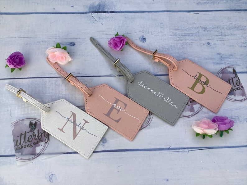 Pu&ograve; includere: Quattro etichette per bagagli personalizzate con nomi e iniziali diversi. Le etichette sono realizzate in un materiale morbido, rosa chiaro e grigio. Le iniziali sono in lettere dorate. Le etichette sono attaccate a un bagaglio con un cinturino in pelle marrone.