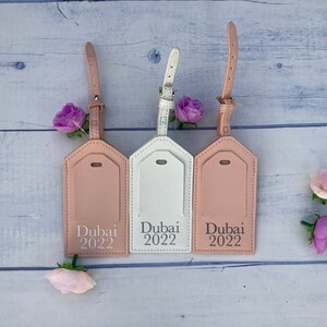 Puede incluir: Tres etiquetas de equipaje con una correa de cuero rosa y un fondo blanco. Las etiquetas son rectangulares con esquinas redondeadas y tienen el texto "Dubai 2022" impreso en ellas.