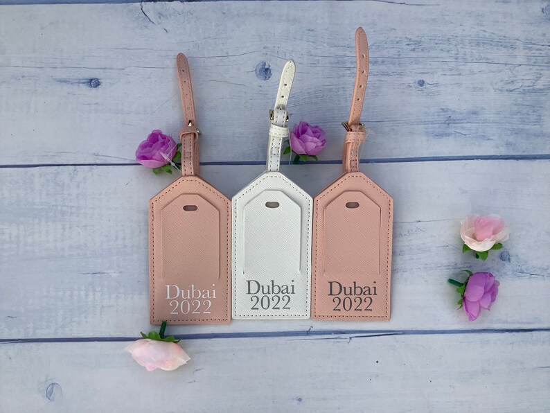 Puede incluir: Tres etiquetas de equipaje con una correa de cuero rosa y un fondo blanco. Las etiquetas son rectangulares con esquinas redondeadas y tienen el texto "Dubai 2022" impreso en ellas.