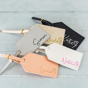 Puede incluir: Cinco etiquetas de equipaje personalizadas con diferentes nombres y un s&iacute;mbolo de coraz&oacute;n. Las etiquetas est&aacute;n hechas de cuero y vienen en diferentes colores, incluyendo gris, beige, negro, blanco y rosa.