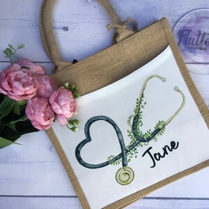 K&ouml;nnte beinhalten: Eine wei&szlig;e Tragetasche mit einem Jute-Au&szlig;enmaterial. Die Tasche hat einen wei&szlig;en Stoffeinsatz mit einem schwarzen und gr&uuml;nen Stethoskop-Design und den Namen "Jane" in Schwarz geschrieben.