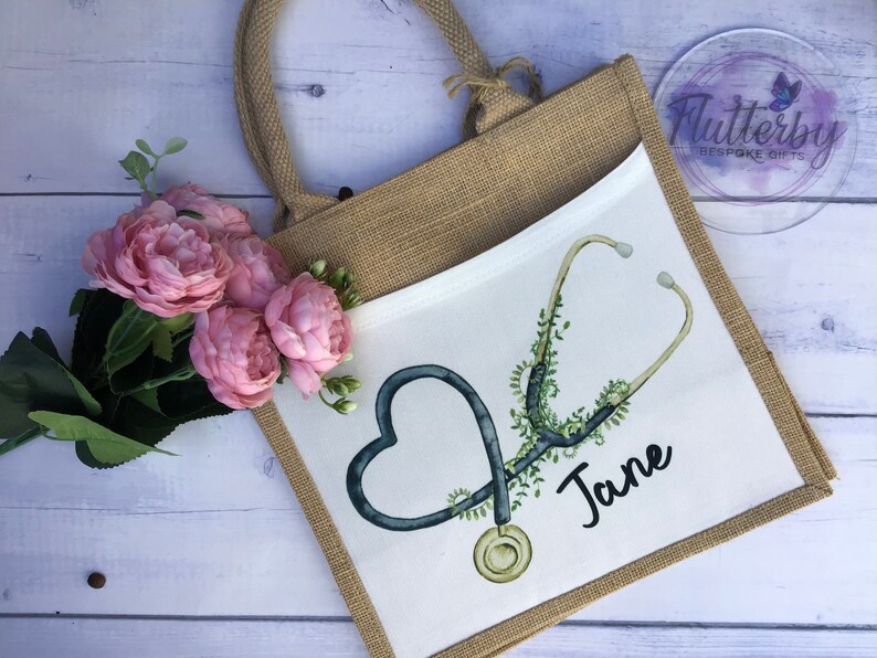 K&ouml;nnte beinhalten: Eine wei&szlig;e Tragetasche mit einem Jute-Au&szlig;enmaterial. Die Tasche hat einen wei&szlig;en Stoffeinsatz mit einem schwarzen und gr&uuml;nen Stethoskop-Design und den Namen "Jane" in Schwarz geschrieben.
