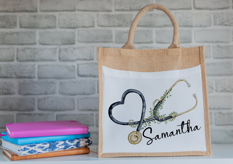 K&ouml;nnte beinhalten: Eine braune Jute-Tragetasche mit einem wei&szlig;en Canvas-Einsatz. Der Canvas-Einsatz zeigt eine schwarz-wei&szlig;e Illustration eines Stethoskops in Herzform mit gr&uuml;nen Bl&auml;ttern. Der Text "Samantha" ist in schwarzer, kursiv geschriebener Schrift unter der Illustration zu sehen.