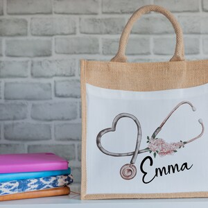K&ouml;nnte beinhalten: Eine nat&uuml;rliche Jute-Tragetasche mit einem wei&szlig;en Canvas-Einsatz, der ein Stethoskop in Herzform mit rosa Blumen und dem Namen "Emma" zeigt.