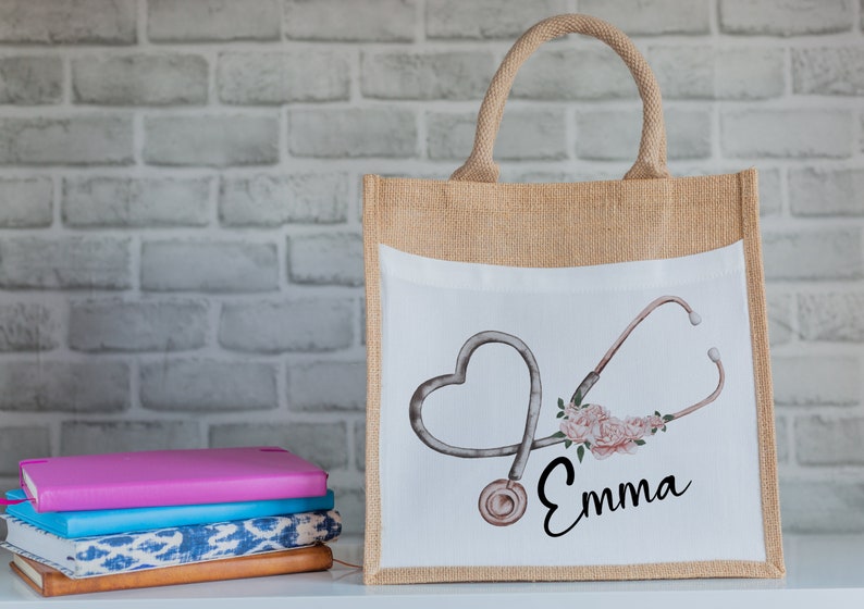 K&ouml;nnte beinhalten: Eine nat&uuml;rliche Jute-Tragetasche mit einem wei&szlig;en Canvas-Einsatz, der ein Stethoskop in Herzform mit rosa Blumen und dem Namen "Emma" zeigt.