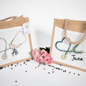 K&ouml;nnte beinhalten: Zwei personalisierte Jute-Tragetaschen mit wei&szlig;en Stoffeins&auml;tzen. Die Taschen zeigen ein Stethoskop mit einem Herzen und einem Blumendesign. Eine Tasche sagt "Ali" und die andere sagt "Jane".