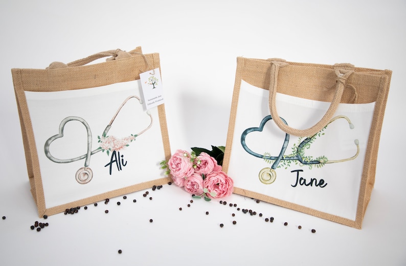 K&ouml;nnte beinhalten: Zwei personalisierte Jute-Tragetaschen mit wei&szlig;en Stoffeins&auml;tzen. Die Taschen zeigen ein Stethoskop mit einem Herzen und einem Blumendesign. Eine Tasche sagt "Ali" und die andere sagt "Jane".